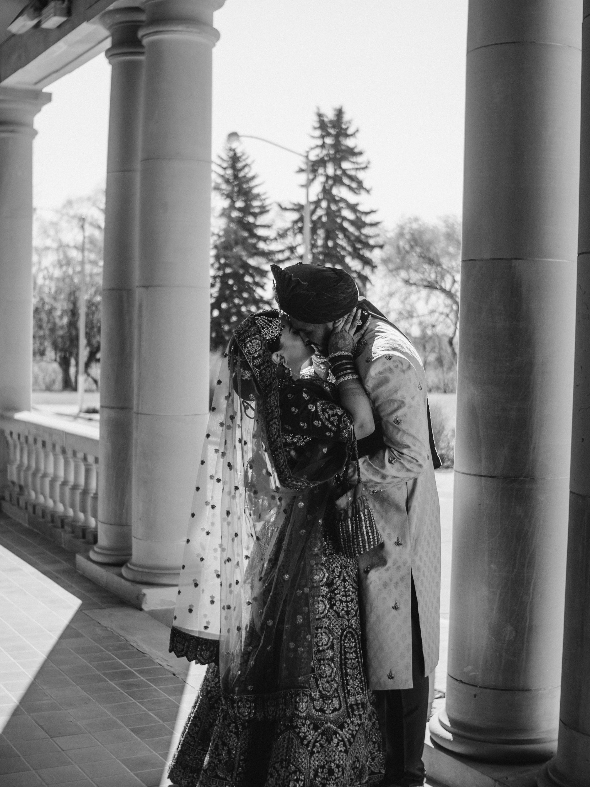 A black and white photo of a bride and groom kissing in front of columns.