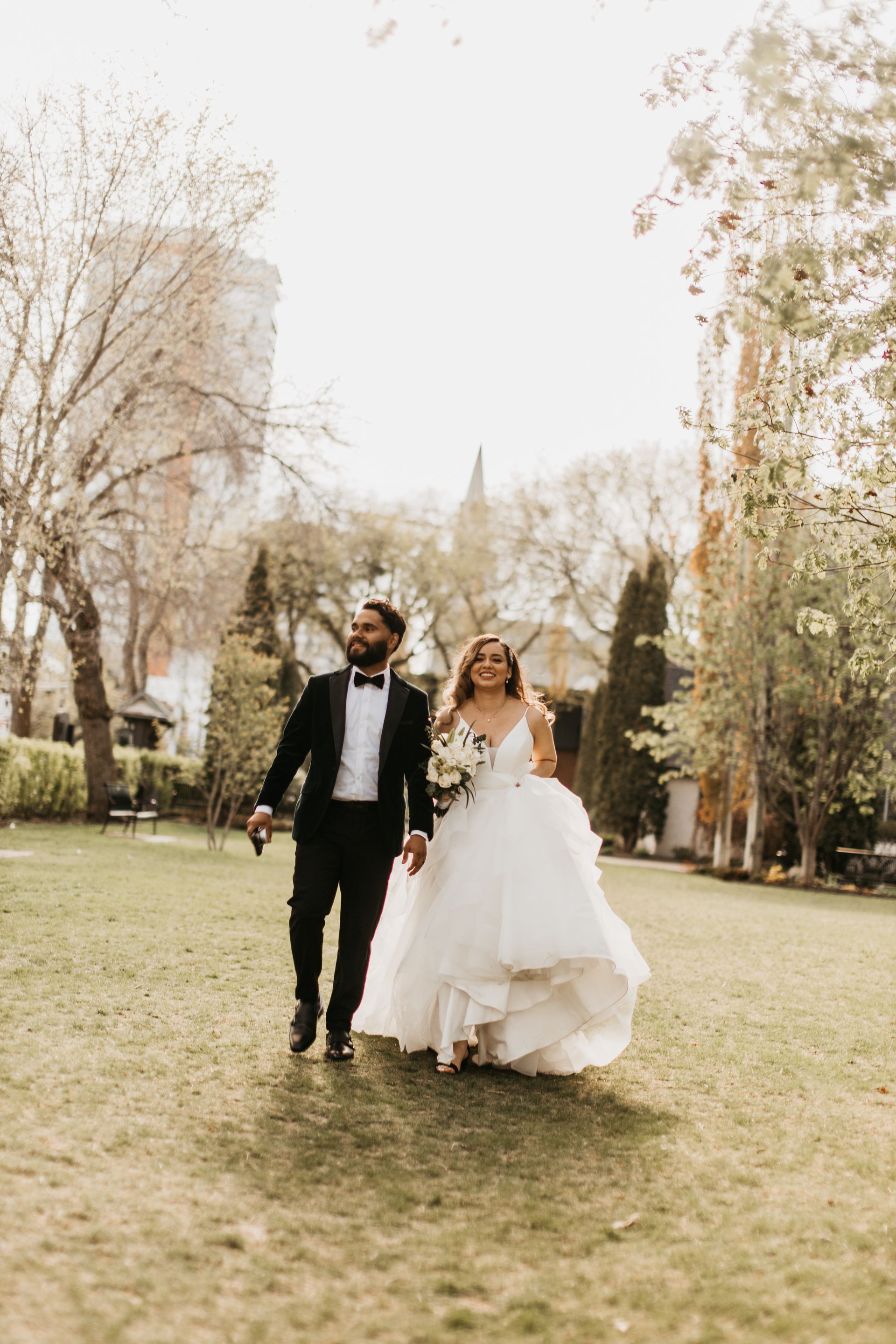A bride and groom are walking through a park holding hands.