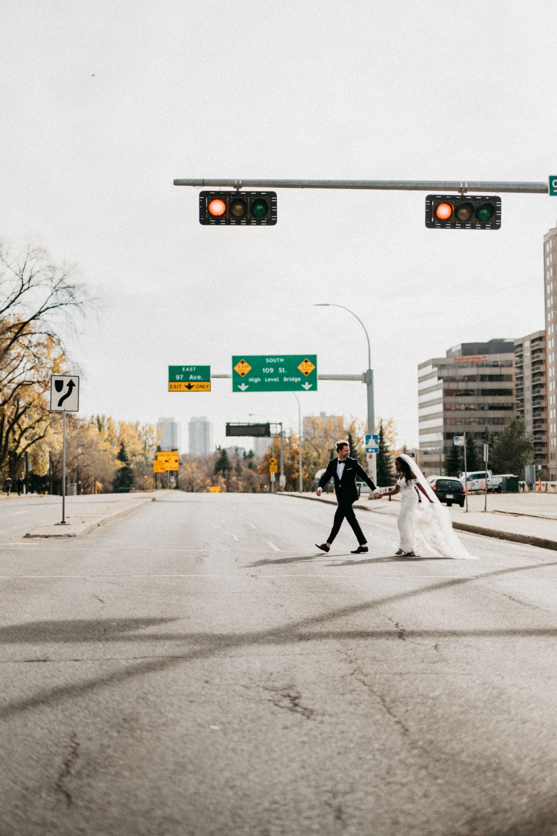 A bride and groom are crossing a street under a traffic light.