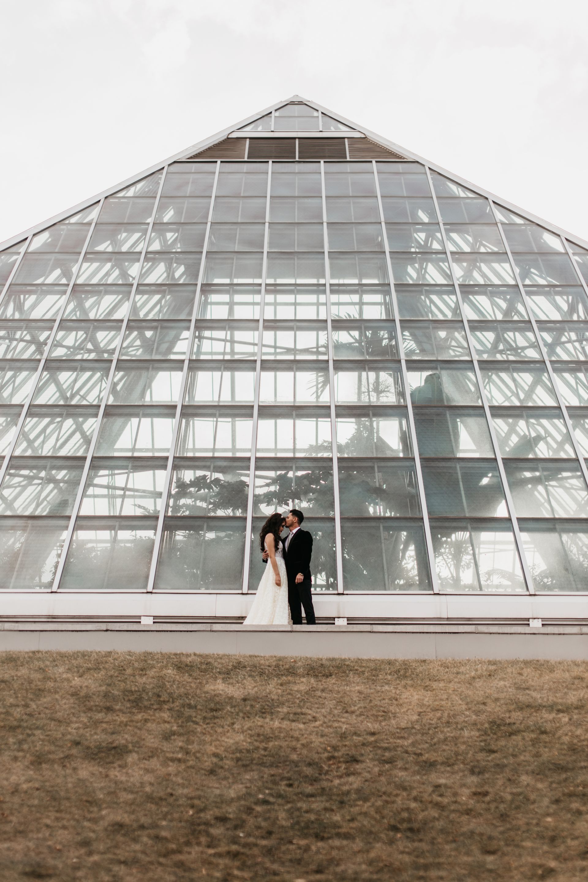 A bride and groom are kissing in front of a large glass building.