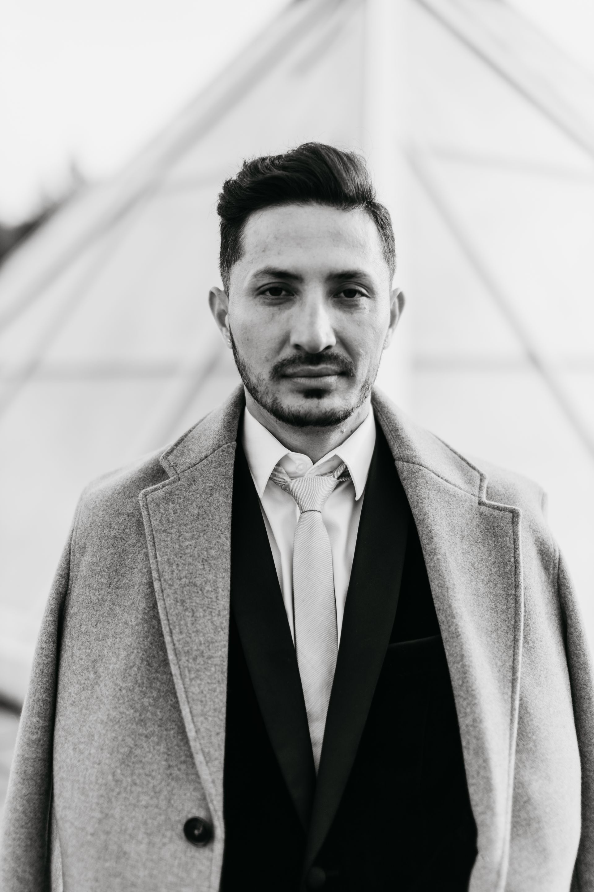A man in a suit and coat is standing in front of a building in a black and white photo.