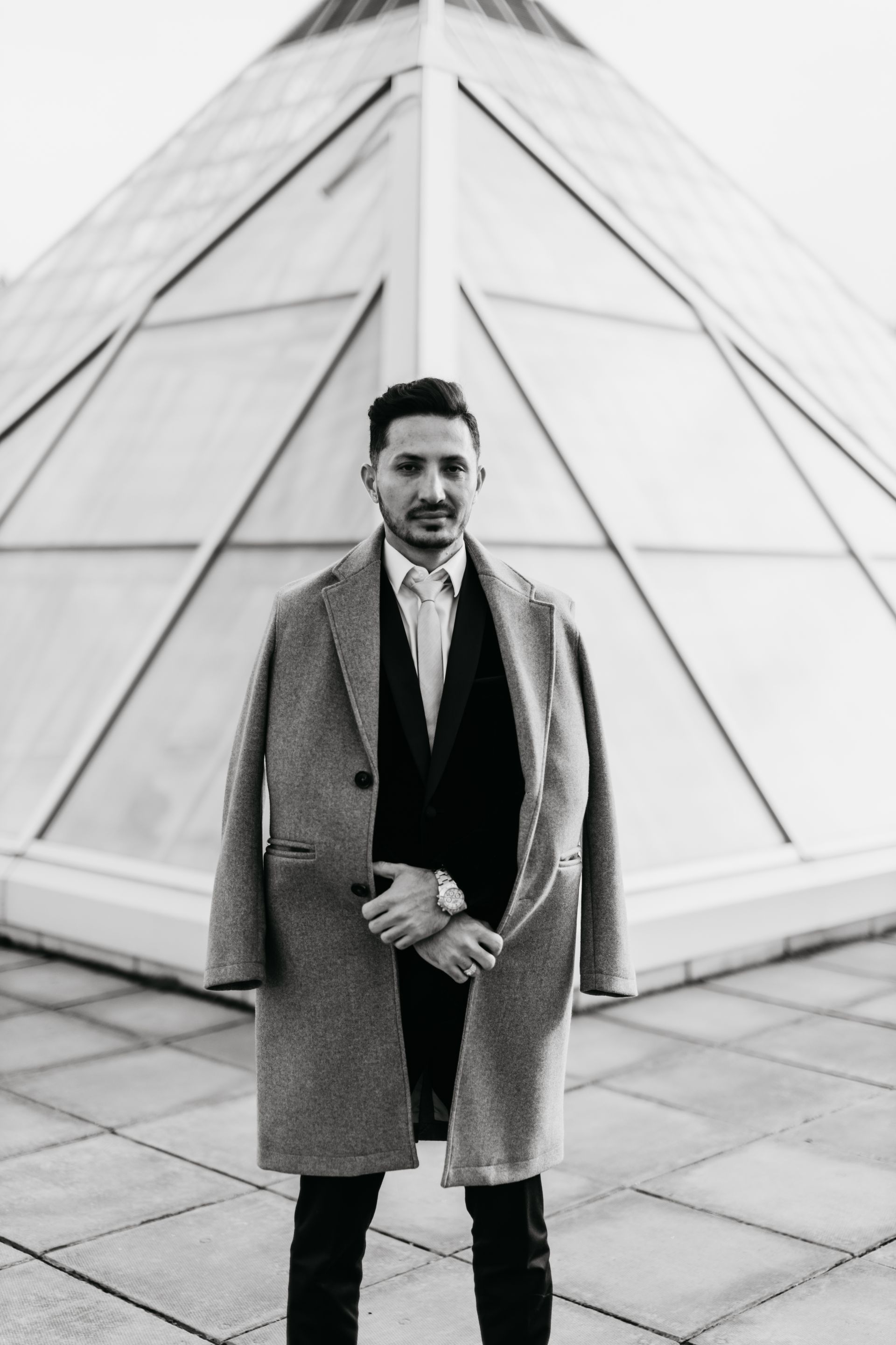 A man in a suit and coat is standing in front of a pyramid.
