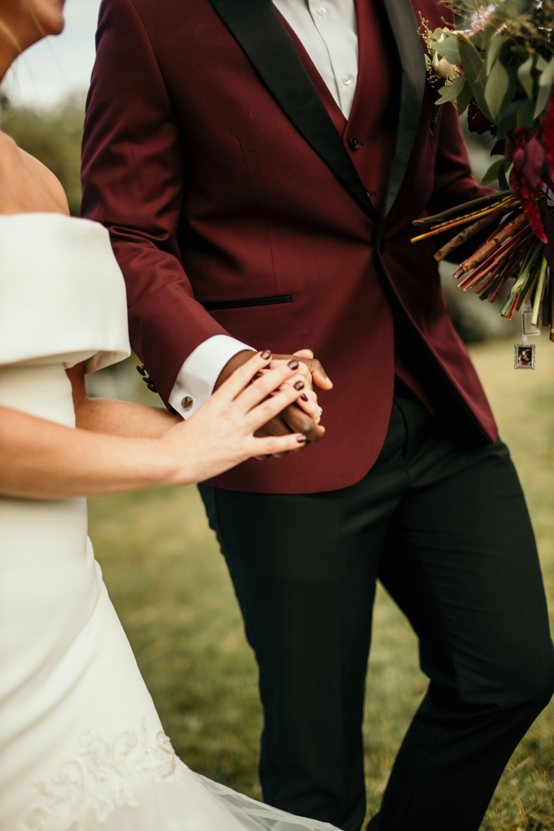 The bride and groom are holding hands and the groom is wearing a maroon tuxedo.