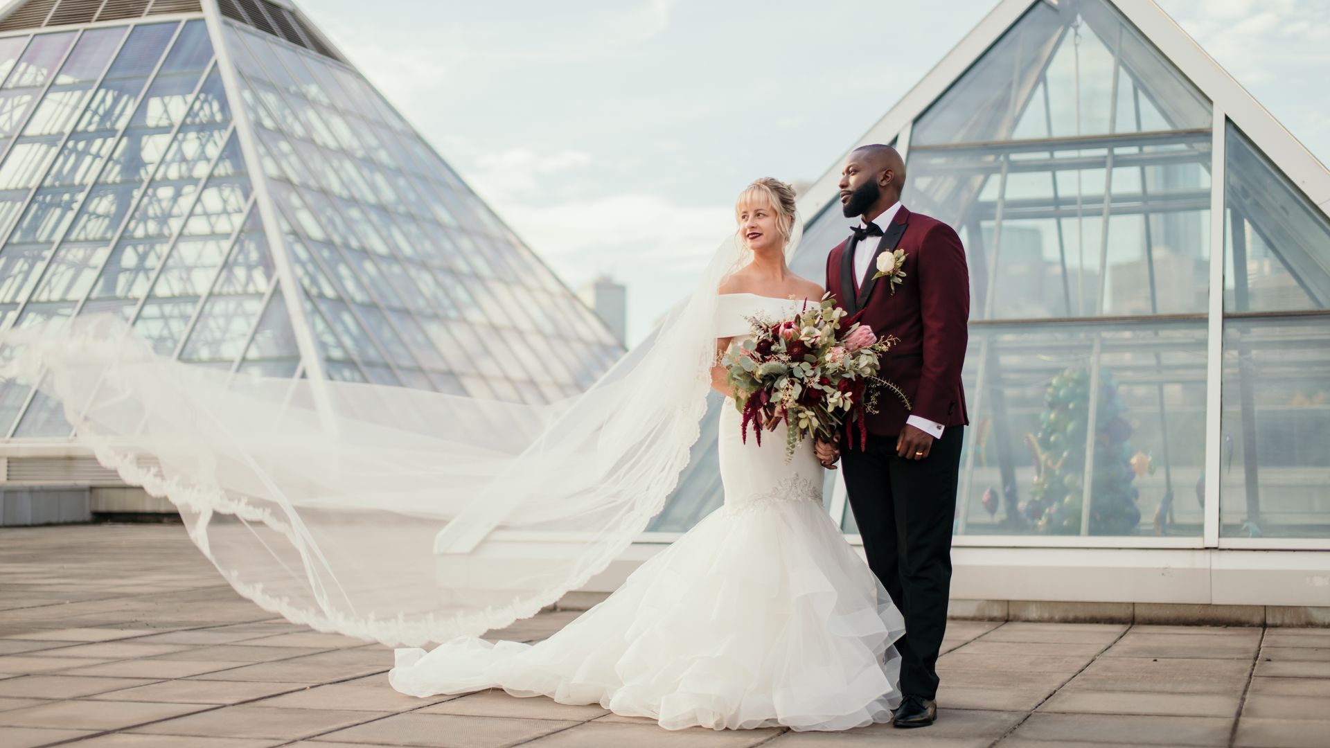 A bride and groom are walking on a rooftop with their veil blowing in the wind.