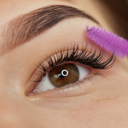 Close-up of eye with long, dark lashes being brushed with a purple spoolie; eyebrow is groomed.