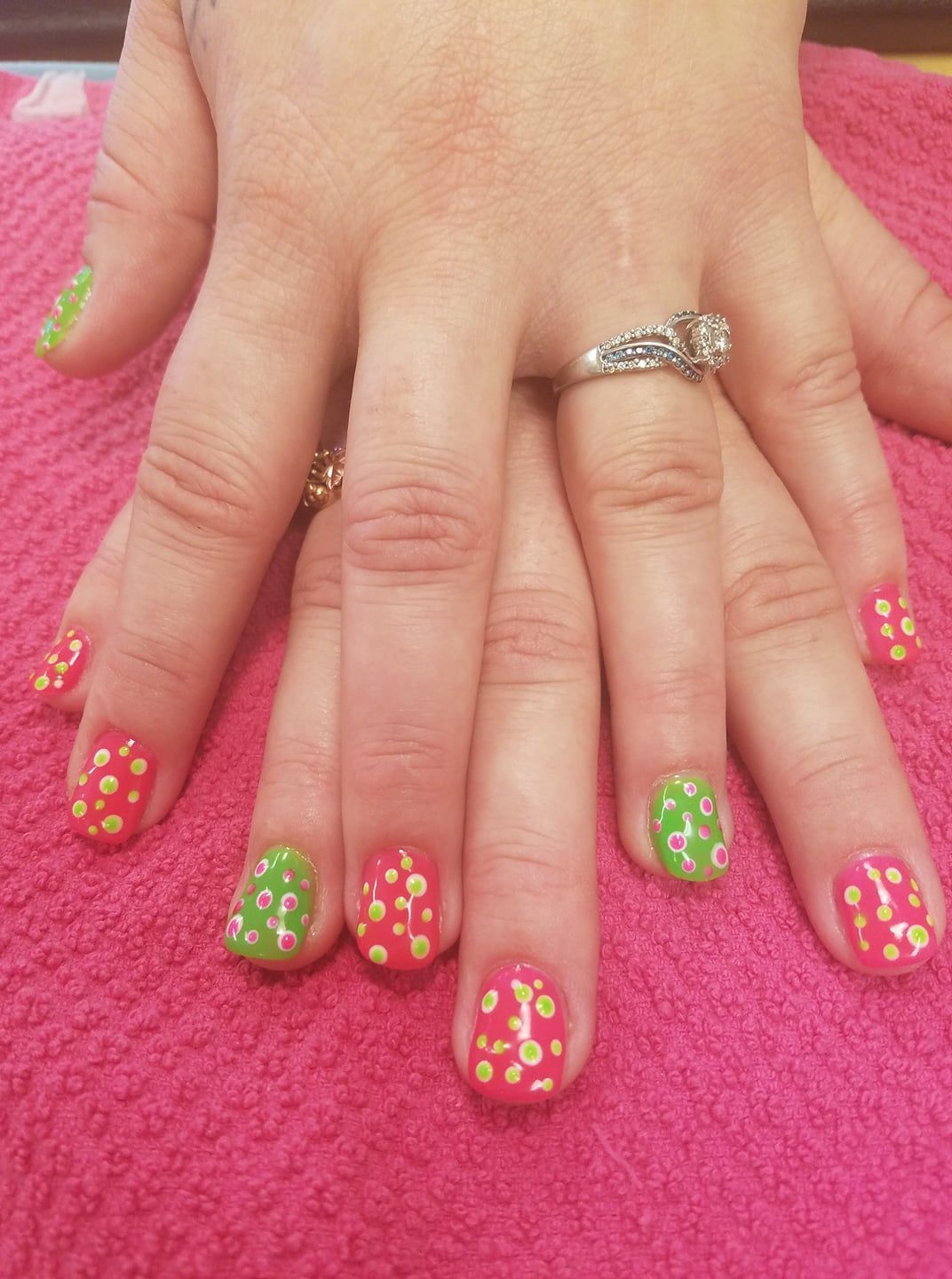 Hands with red and green nails, decorated with yellow polka dots and white flower designs, on a pink towel.