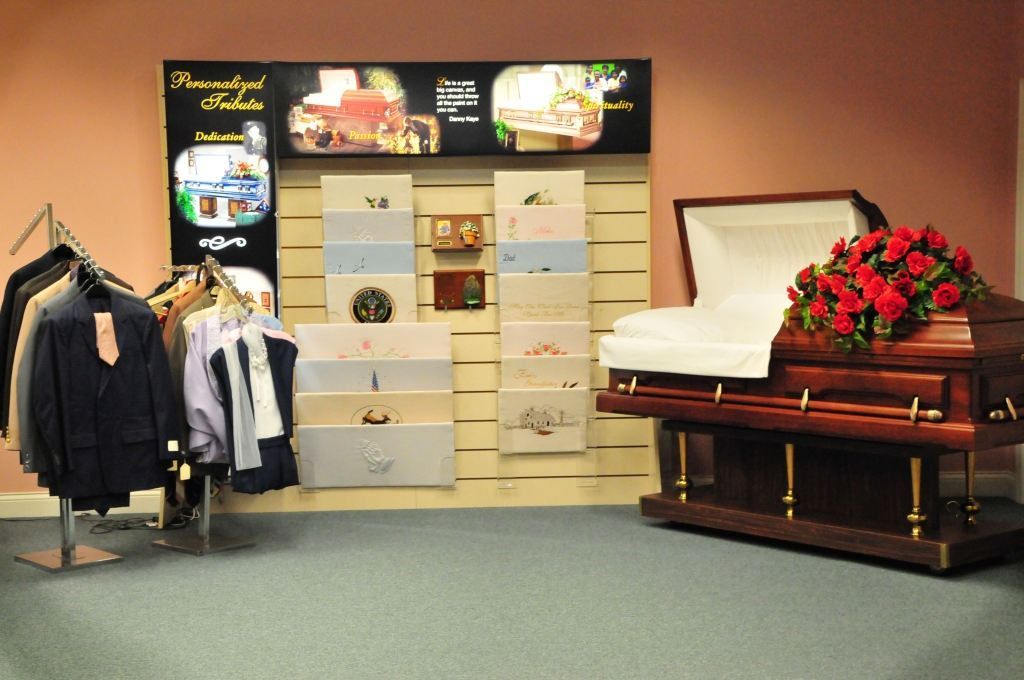 Funeral home display: open casket, clothing rack, urns, and memorial options against a pink wall.