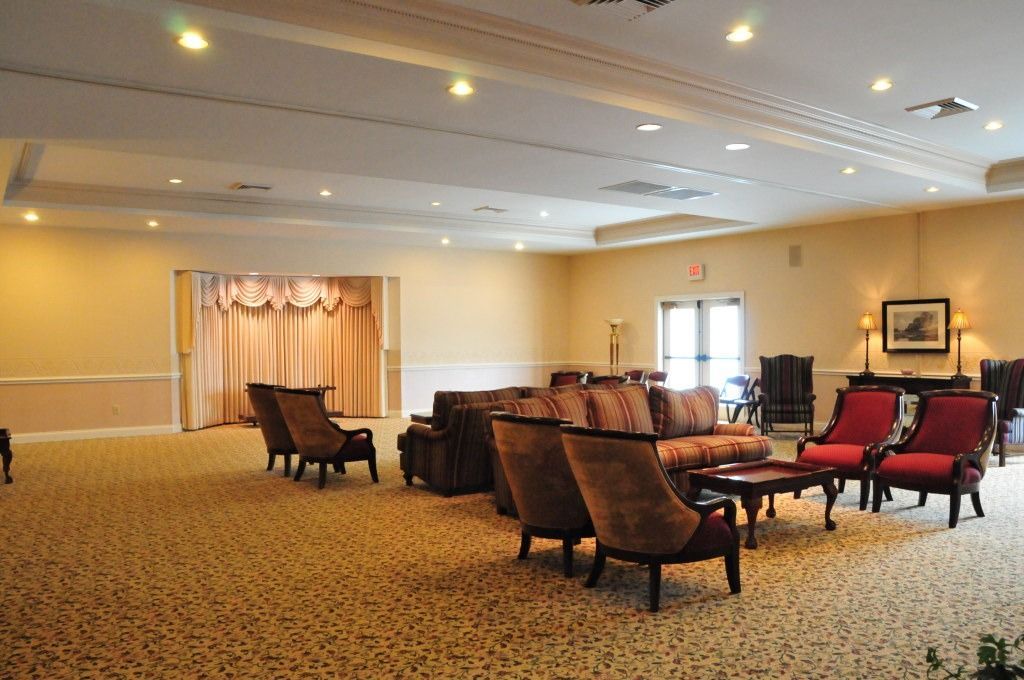 Large, empty room with seating; stage with curtain on the left; carpeted floor and beige walls.