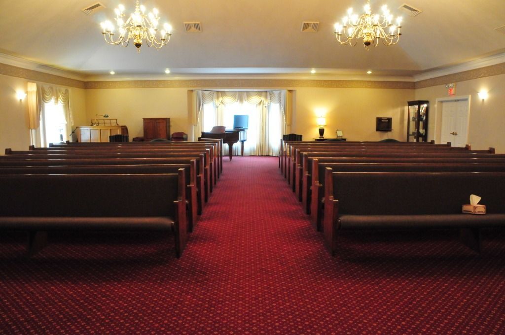 Interior of a funeral home chapel with rows of pews, red carpet, and a piano.