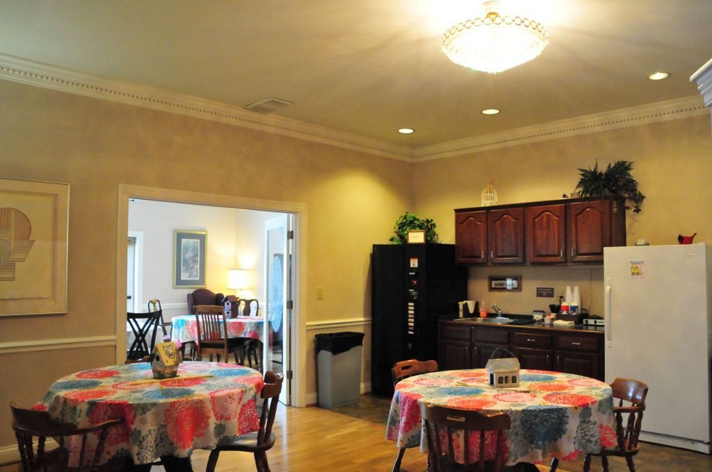 Dining area with round tables, floral tablecloths, kitchen and open doorway to another dining space.