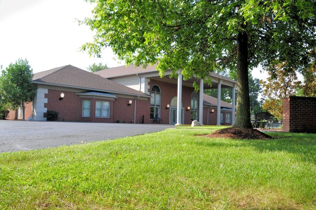 Brick building with columns and a brown roof, next to a tree on a green lawn.