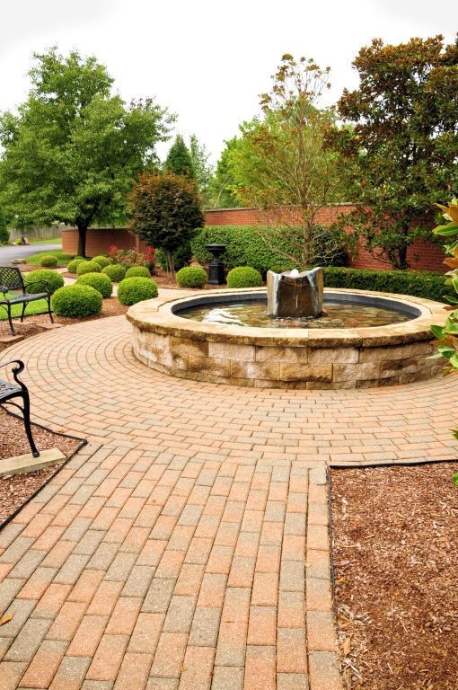 Brick pathway leads to stone fountain in garden, surrounded by shrubs and trees.