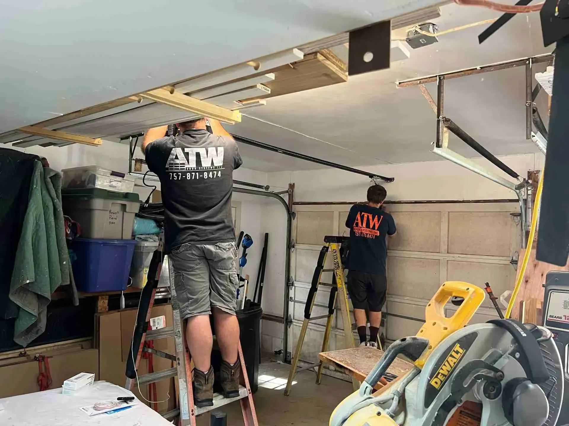 Two men installing a garage door opener, using ladders and tools in a cluttered garage. Two men installing a garage door opener, using ladders and tools in a cluttered garage.