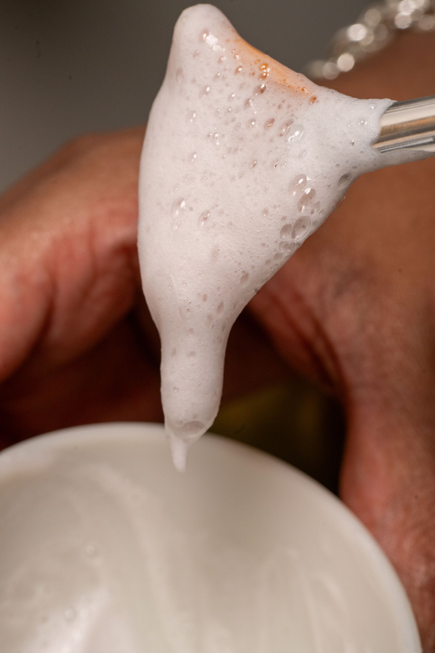 Foamy white substance dripping from a small spatula into a white bowl. Close-up shot, hand holding tools.
