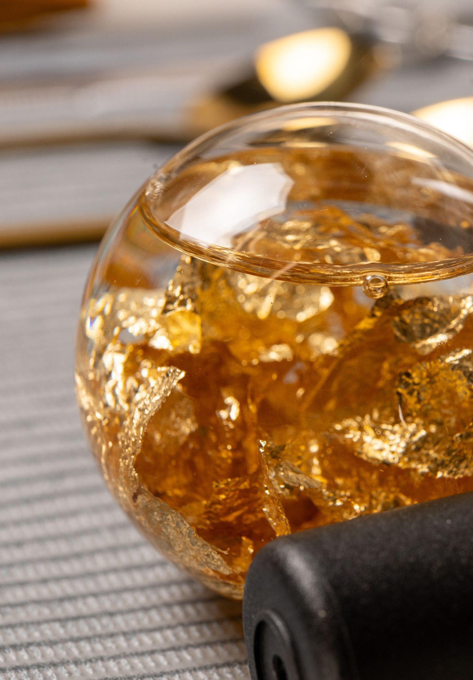 Close-up of a glass orb filled with gold leaf, resting on a striped surface. Black object in the foreground, gold utensils in the blurred background.