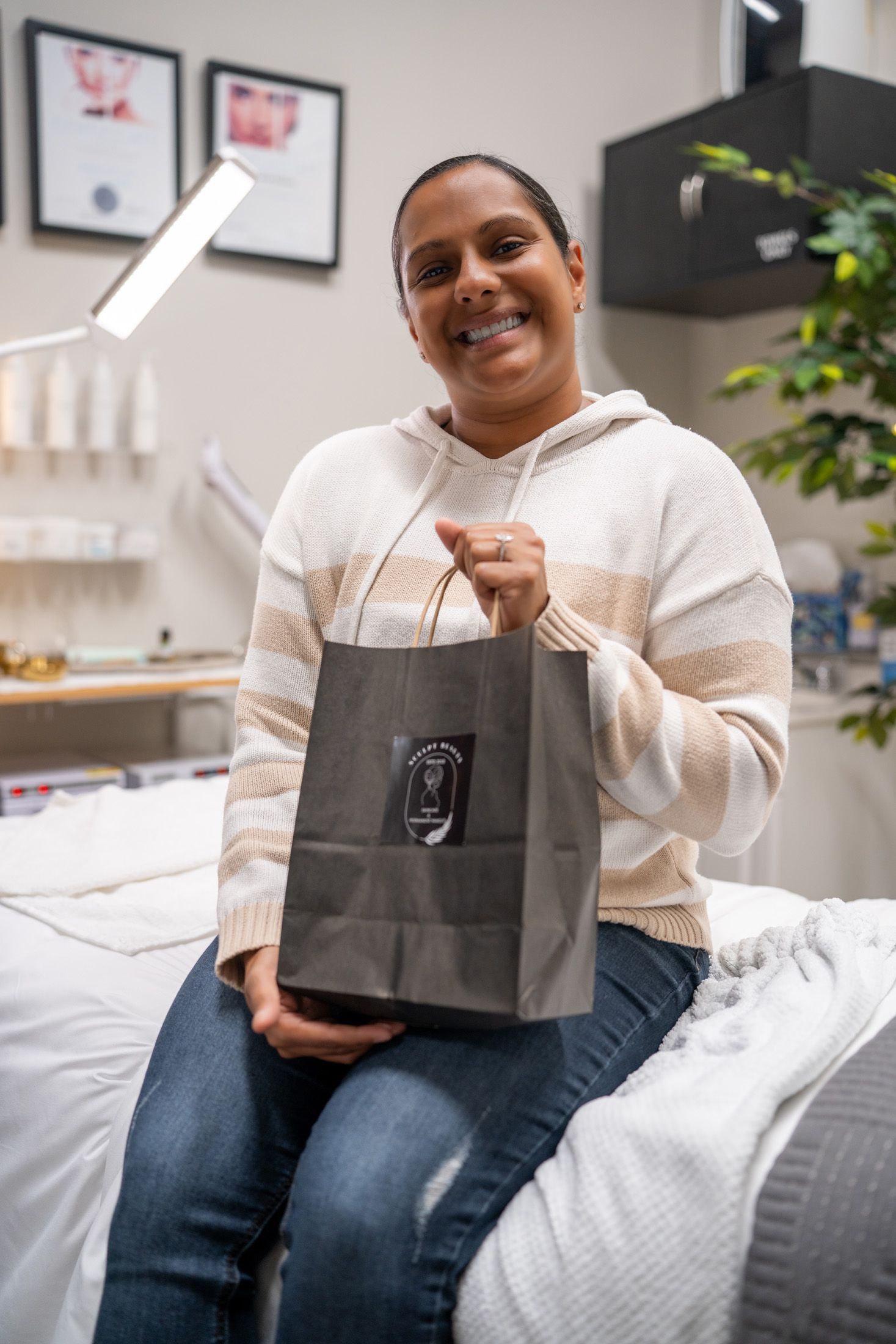 Woman holding a gift bag, smiling. Sitting on a bed in a light-filled spa setting.