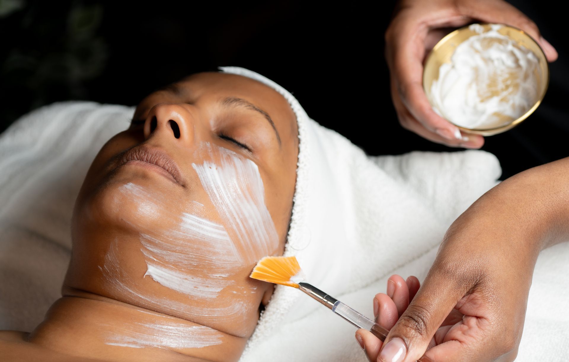 Woman receiving facial treatment, creamy mask being applied with a brush in a spa setting.