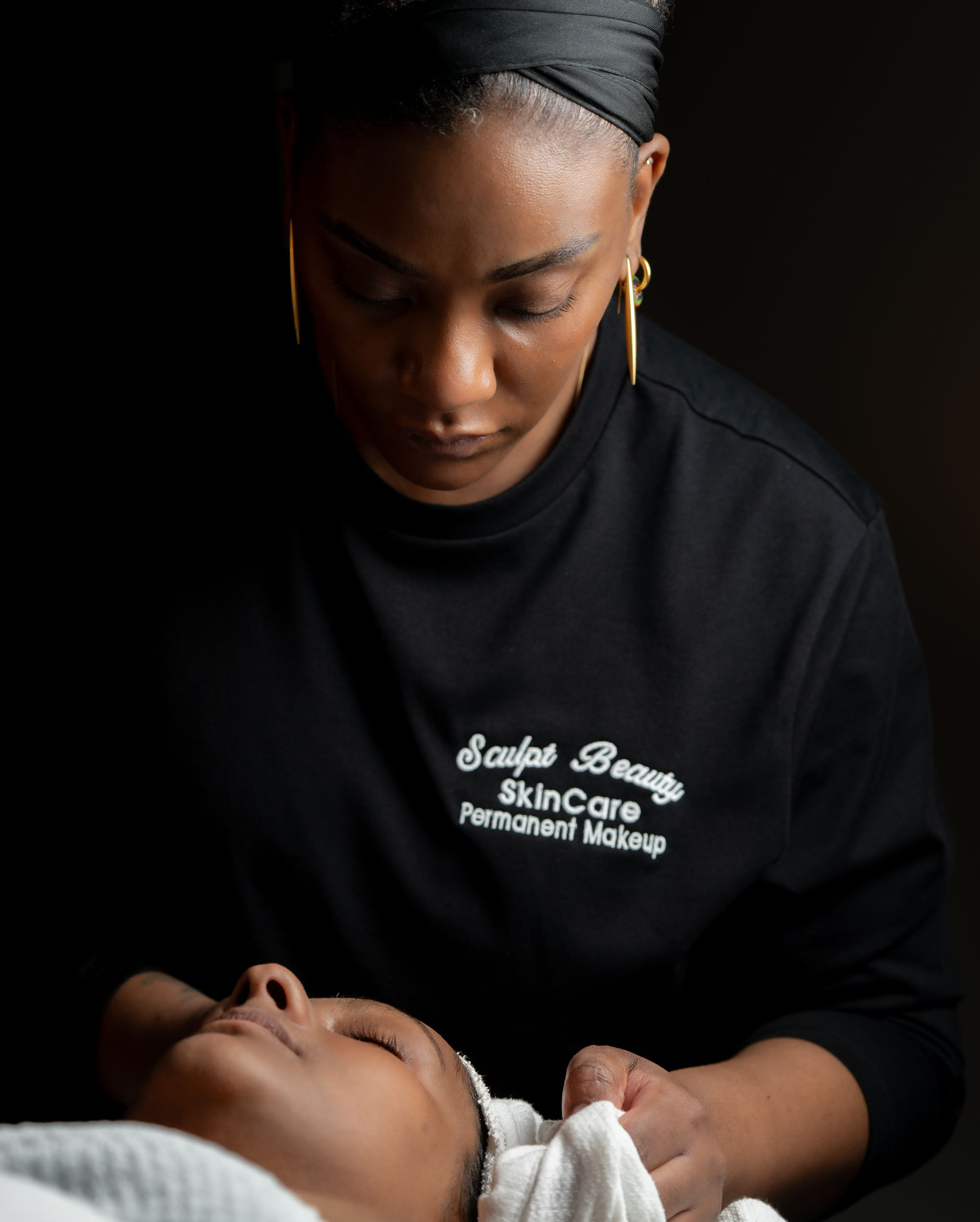 Woman performing a facial, looking down at a client; dimly lit. Black shirt with business logo.