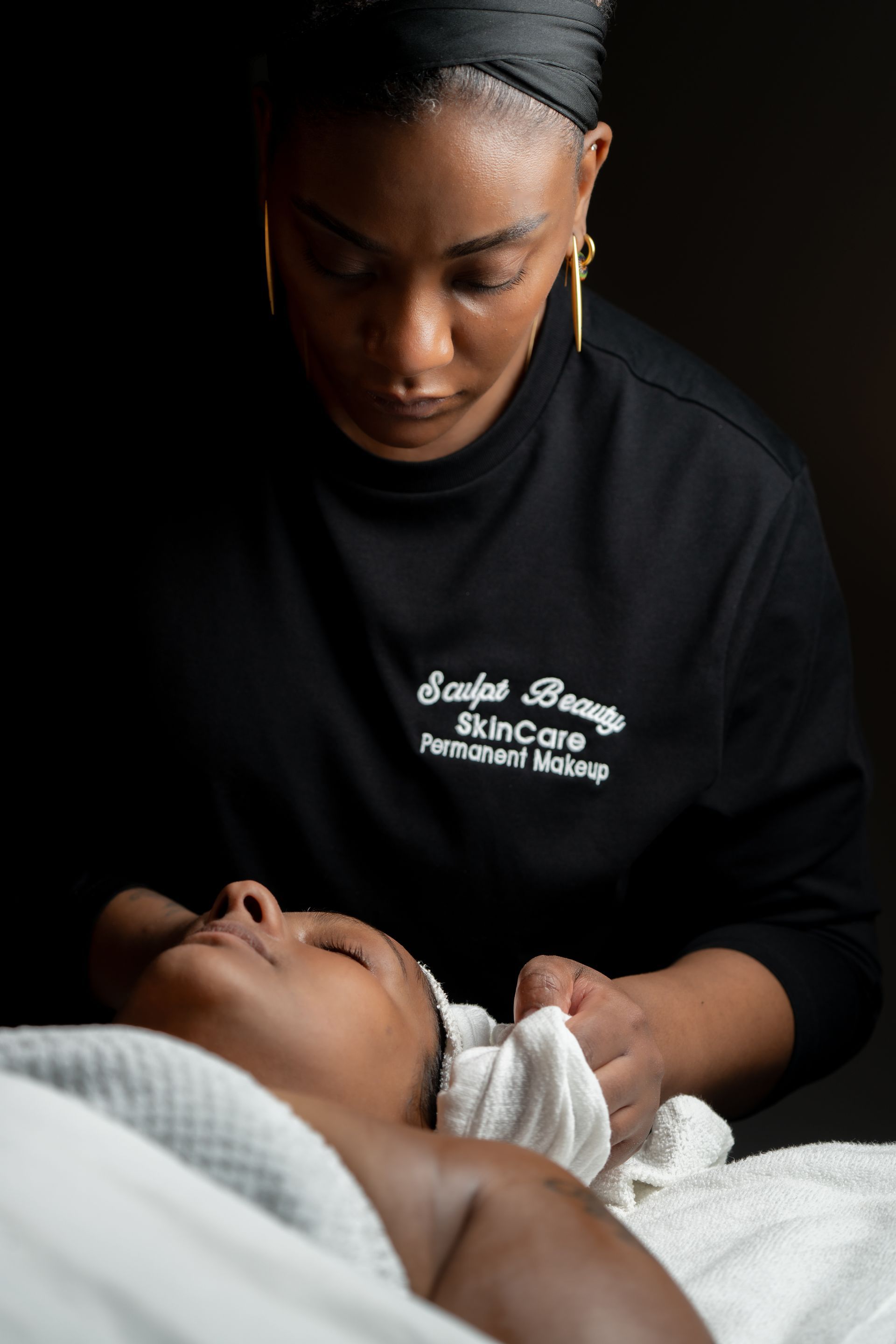 Woman giving facial treatment. She holds a warm cloth, focusing on a client's face. Dark background.