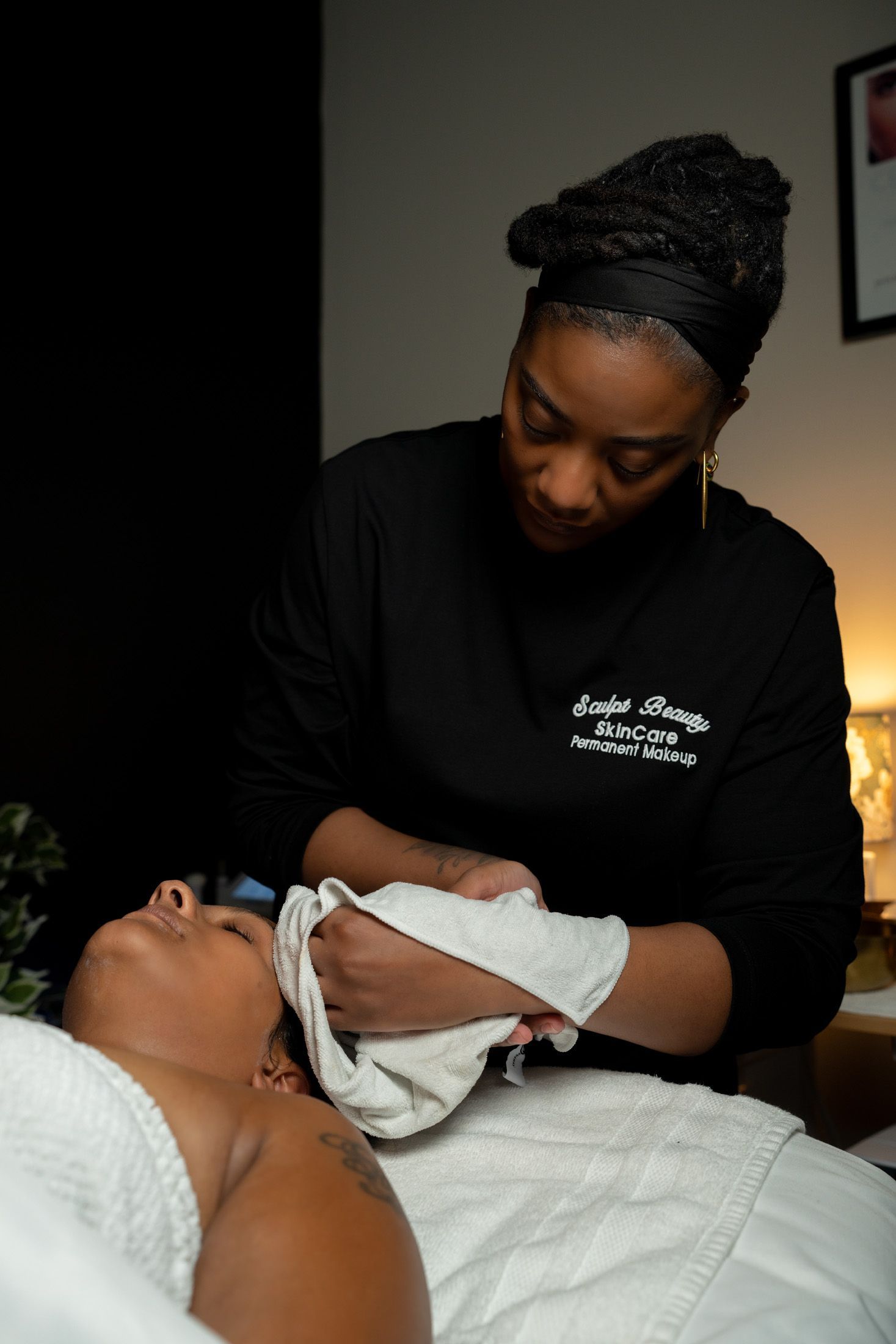 Woman giving facial, wiping face with a towel. Black shirt, warm lighting, cream linens.
