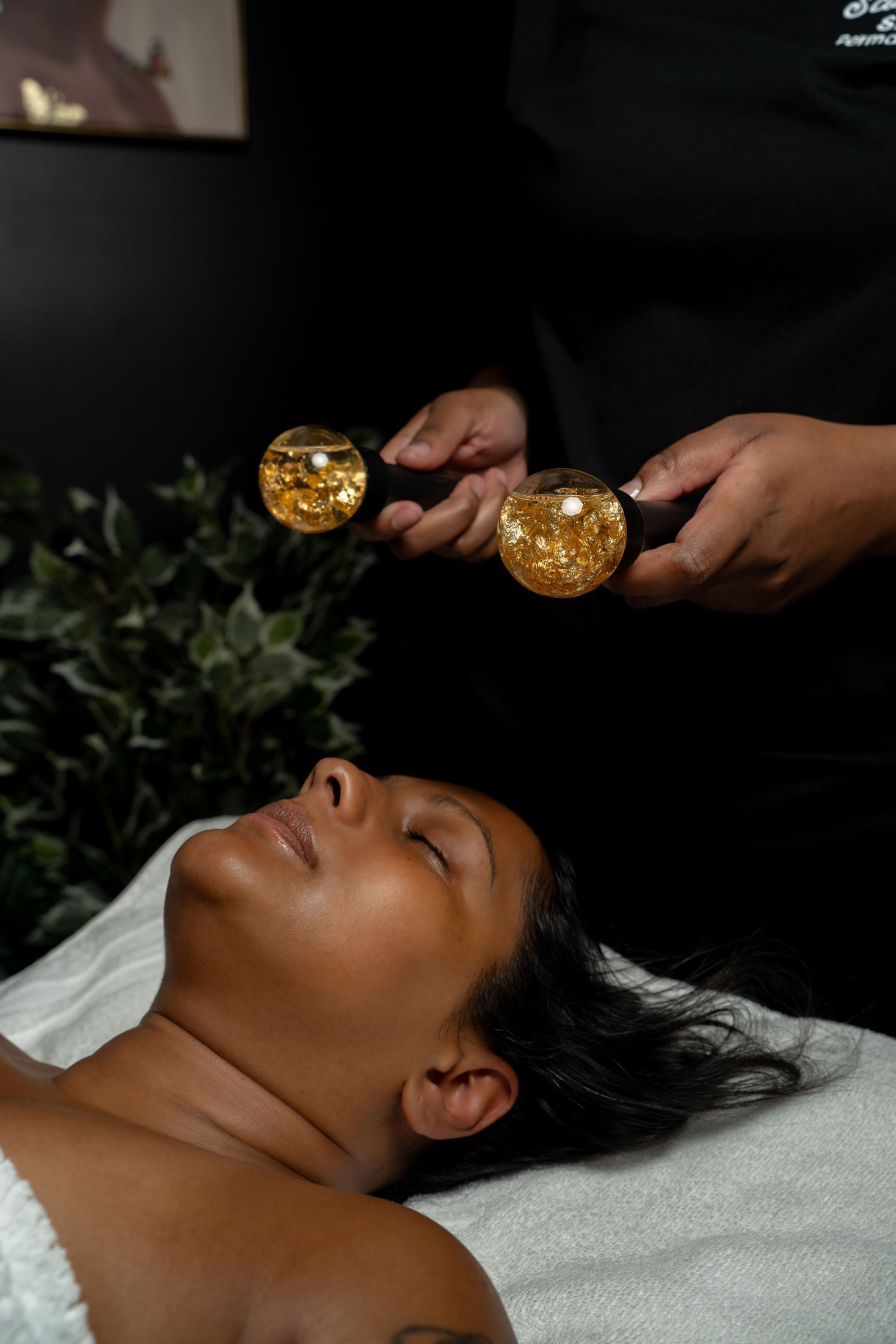 Woman receiving facial treatment; therapist holding two gold globes above her face in a spa setting.