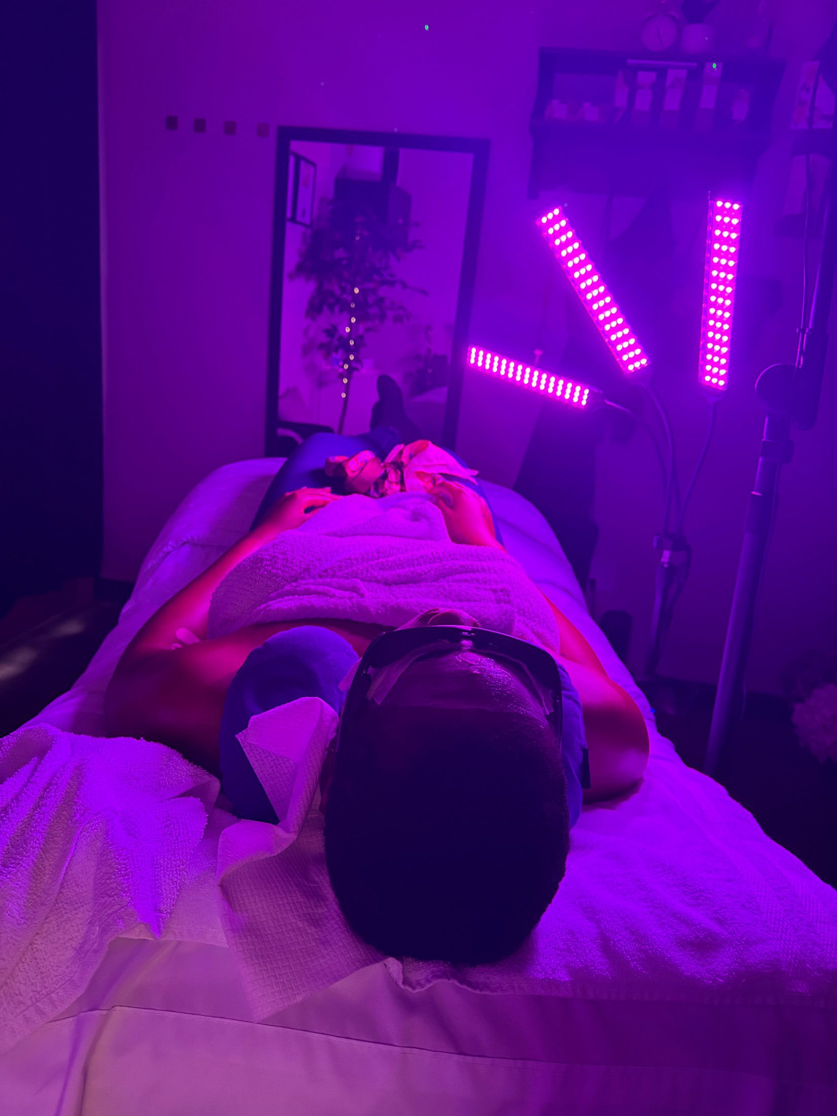 Person receiving light therapy, illuminated by pinkish-purple lights, lying on a white bed.