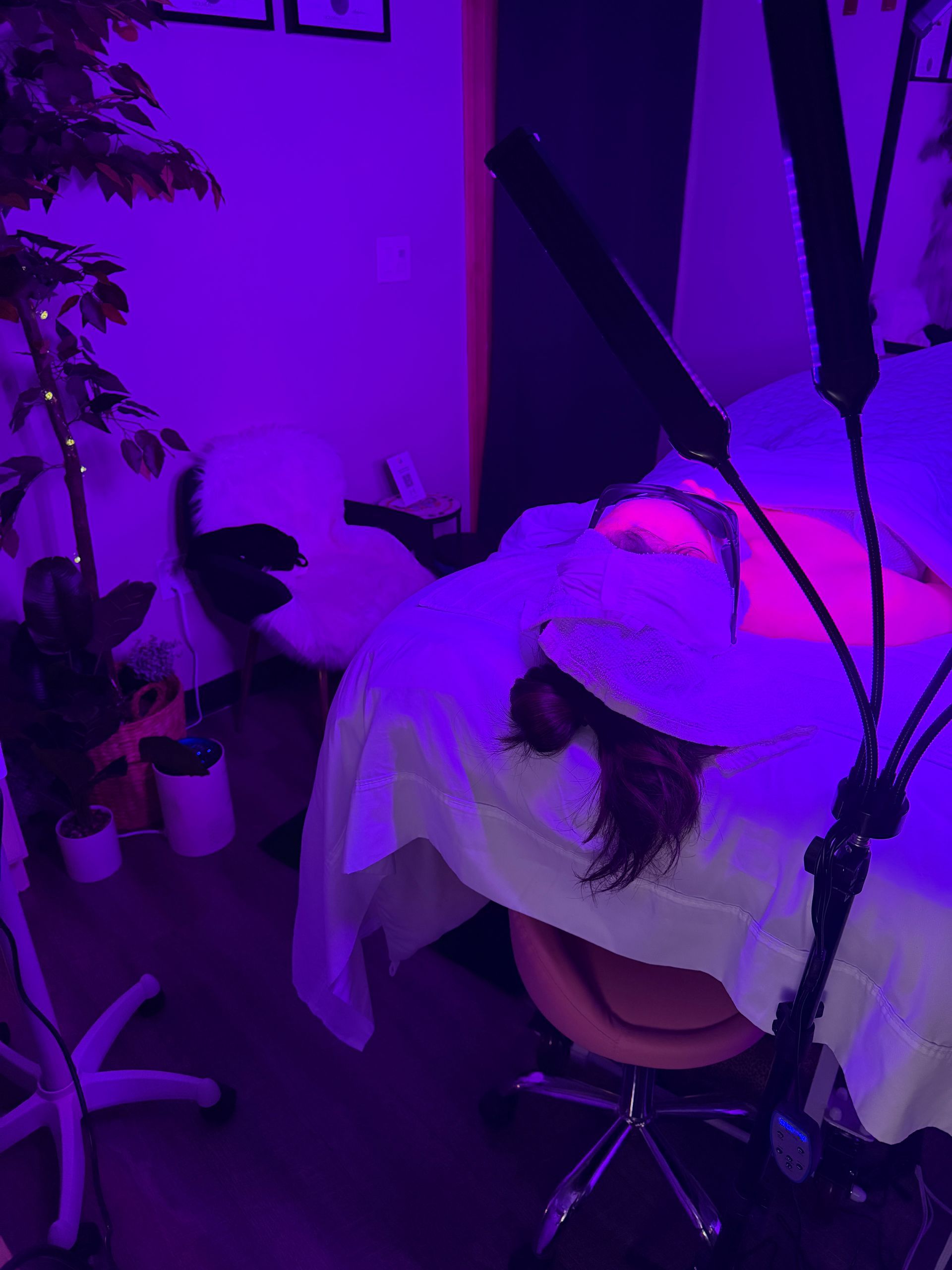 Aesthetician's room with a client on a massage table under a purple glow, lit by a ring light. Plants and a stool are also present.