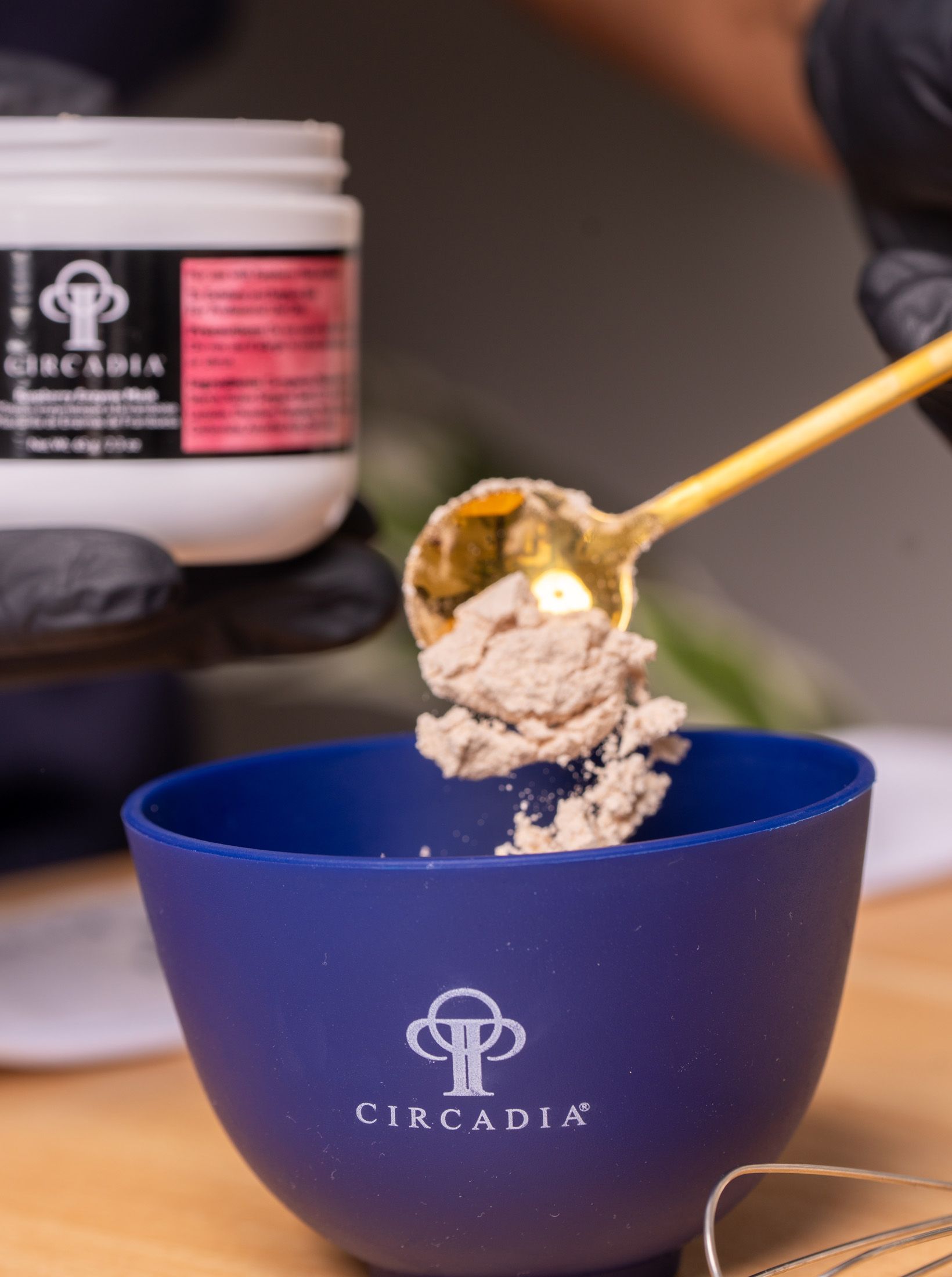 A person wearing black gloves scoops cream from a jar into a blue bowl. The jar is labeled Circadia.