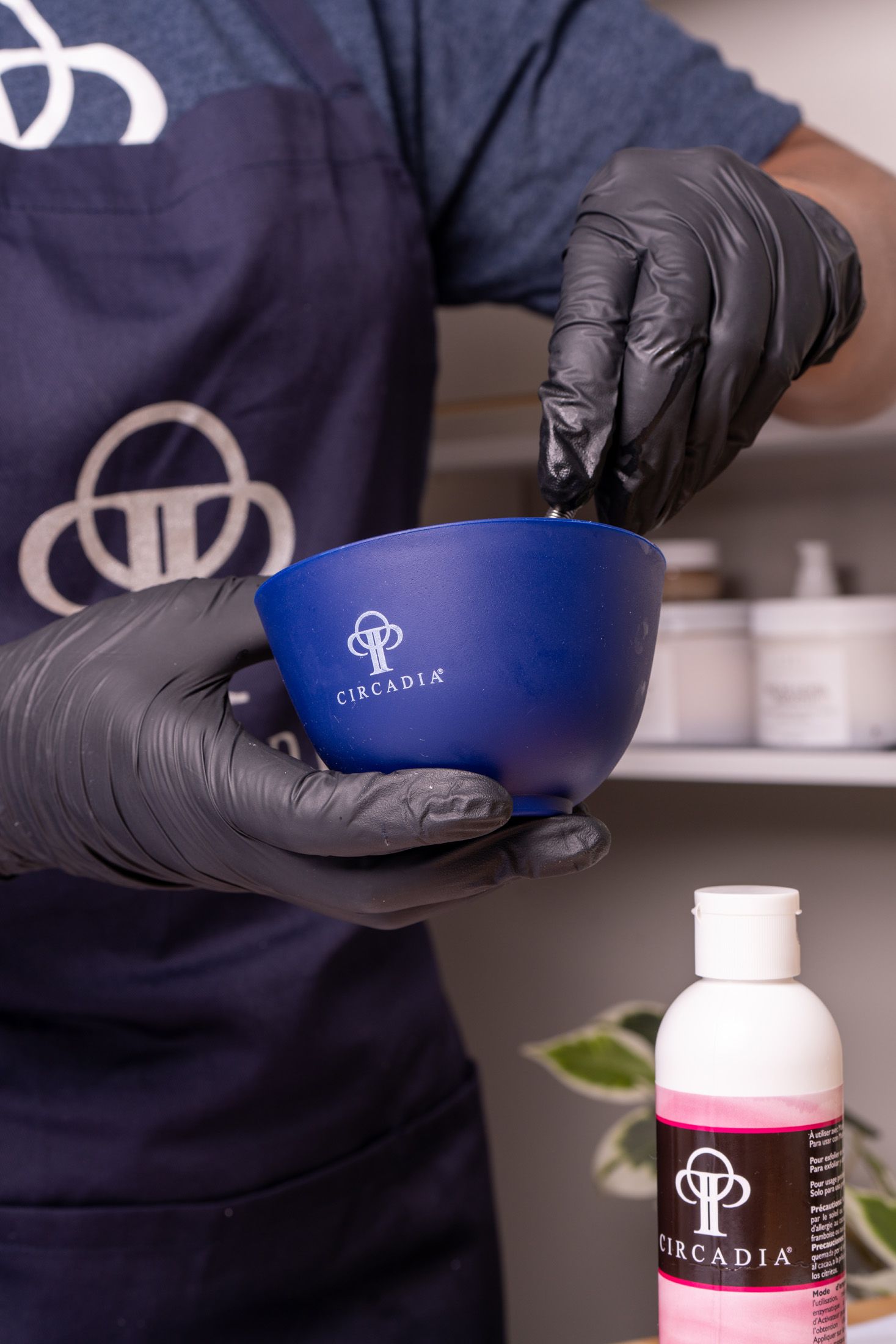 Person with gloved hands mixing a substance in a blue bowl, near a pink liquid bottle.