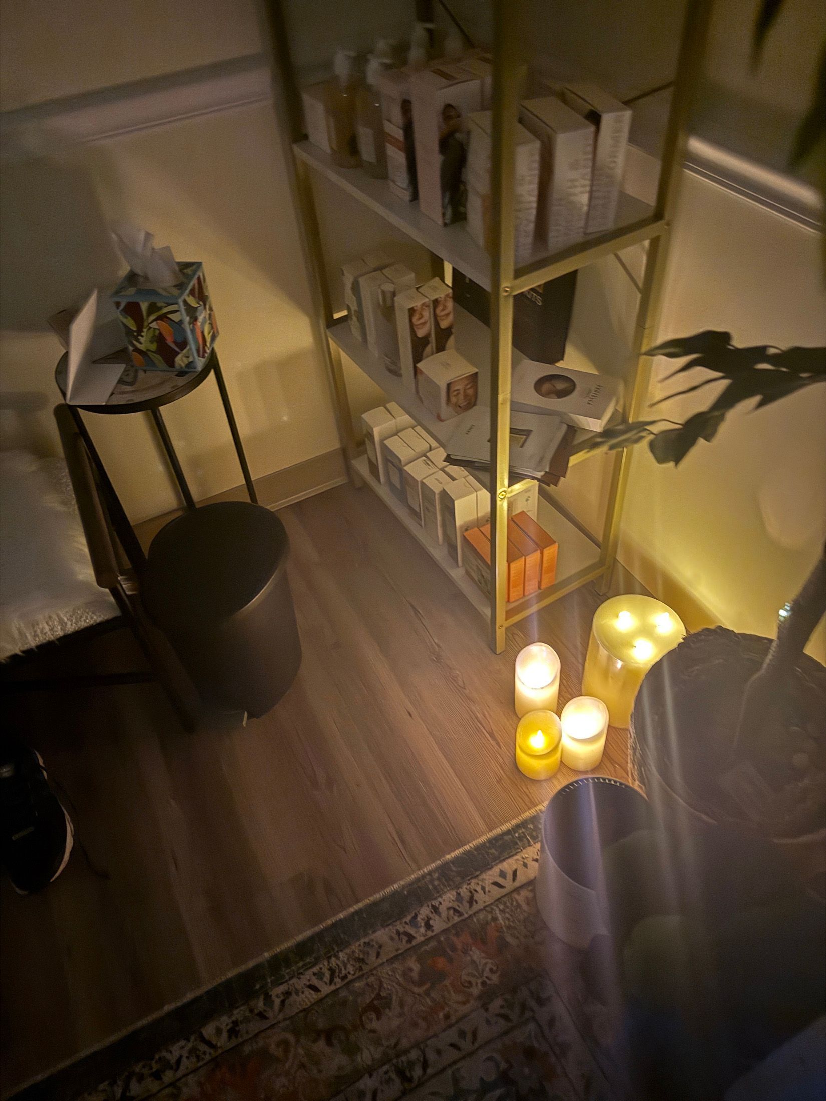 Cozy corner with lit candles, gold shelving unit displaying products, small table, and potted plant on wooden floor.