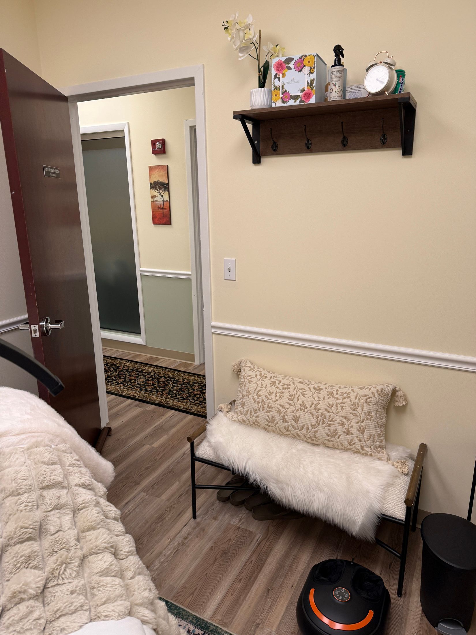 A room with beige walls, a bench with a fur throw, a shelf with decor, and a doorway.