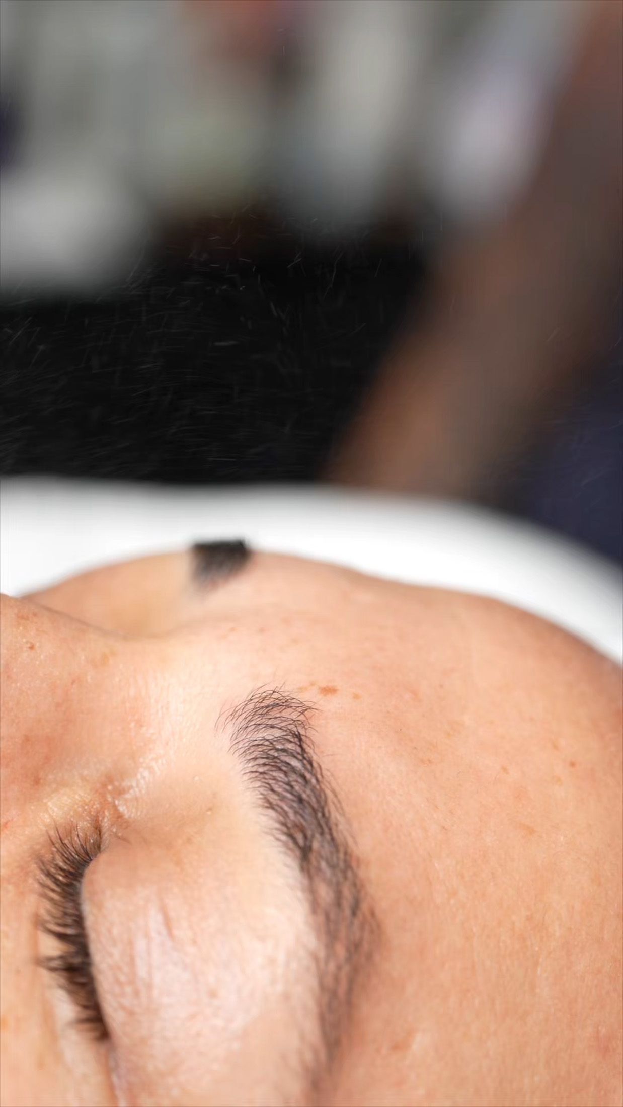 Close-up of a person's closed eye, eyebrow, and forehead. A dark dot is visible on the forehead.