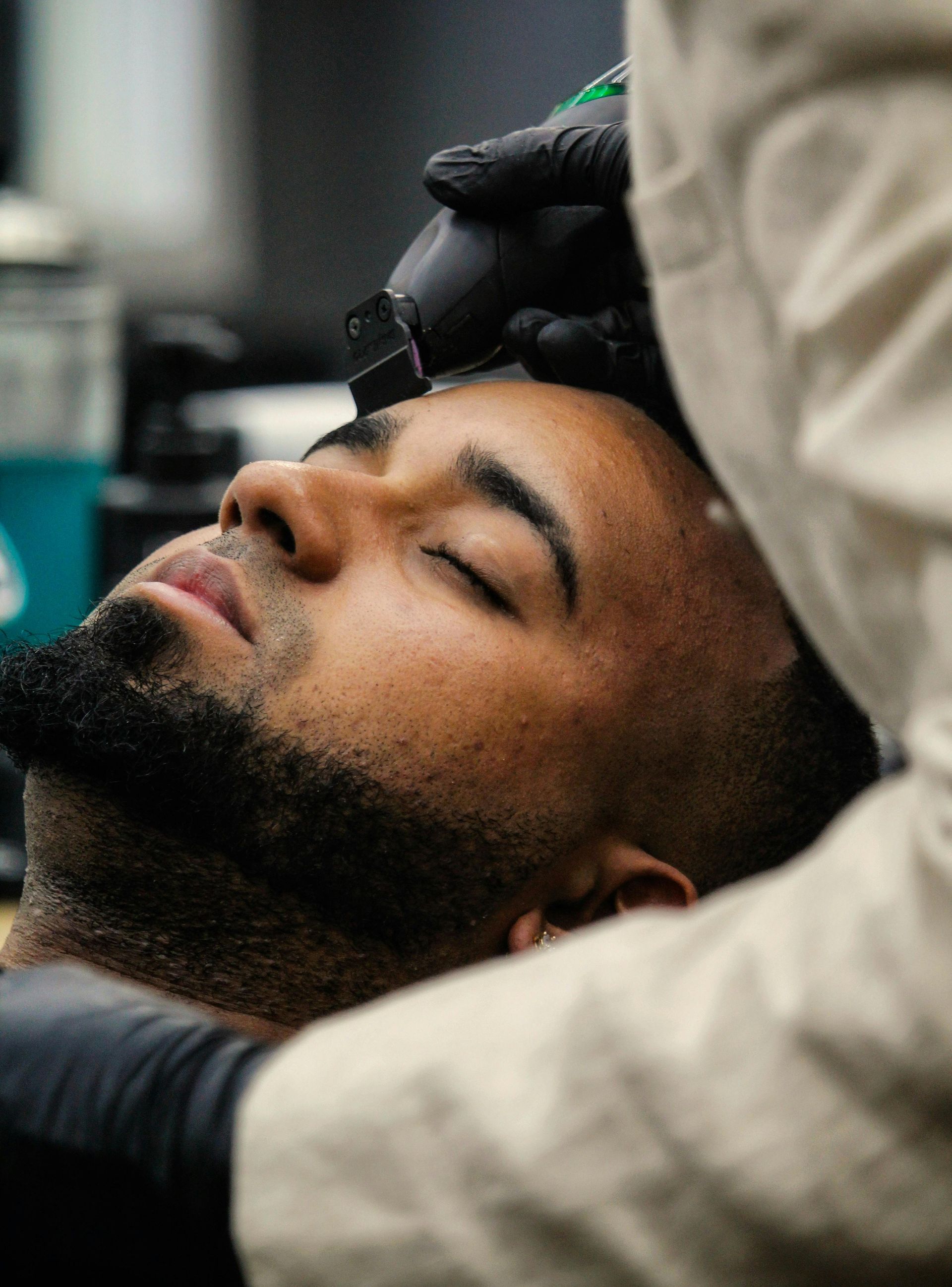 Barber trimming a person's hairline with clippers; face relaxed, eyes closed; black gloves, light-colored smock.