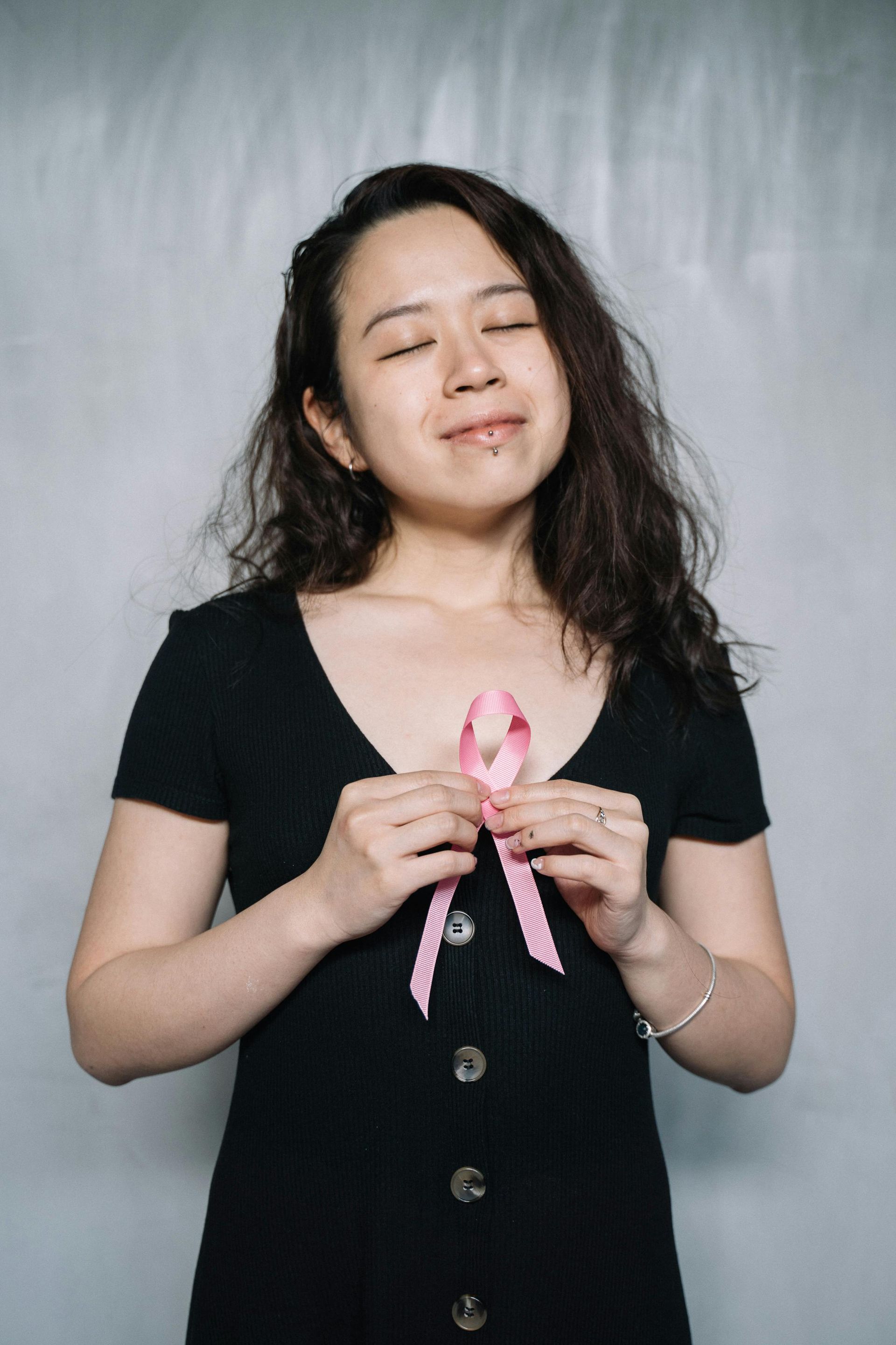 Woman holding pink ribbon, eyes closed, against a gray backdrop.