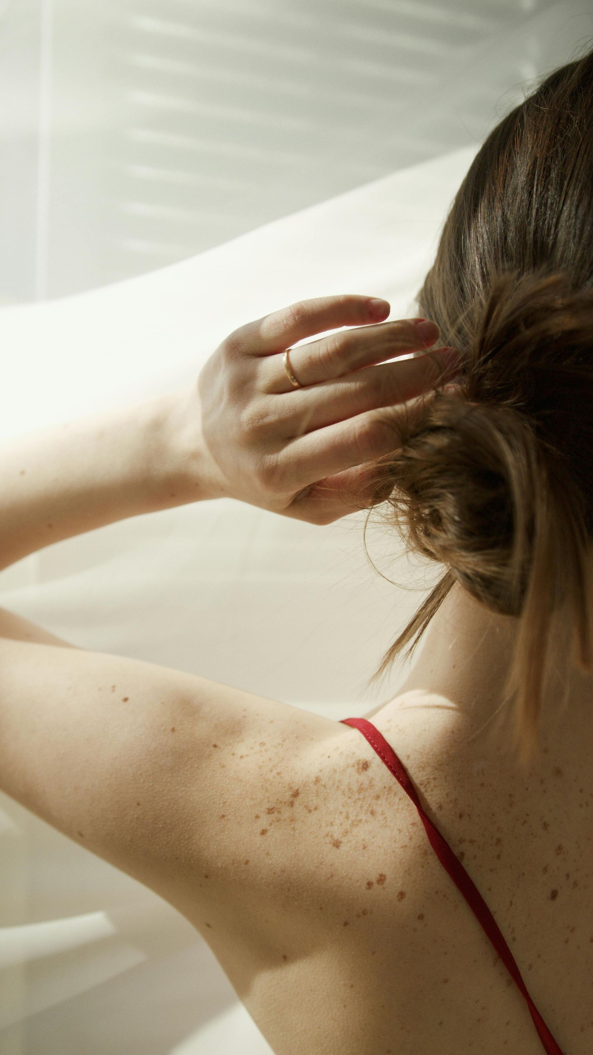 Woman's arm and shoulder, red strap top, hand in hair. Sunny, bright background.