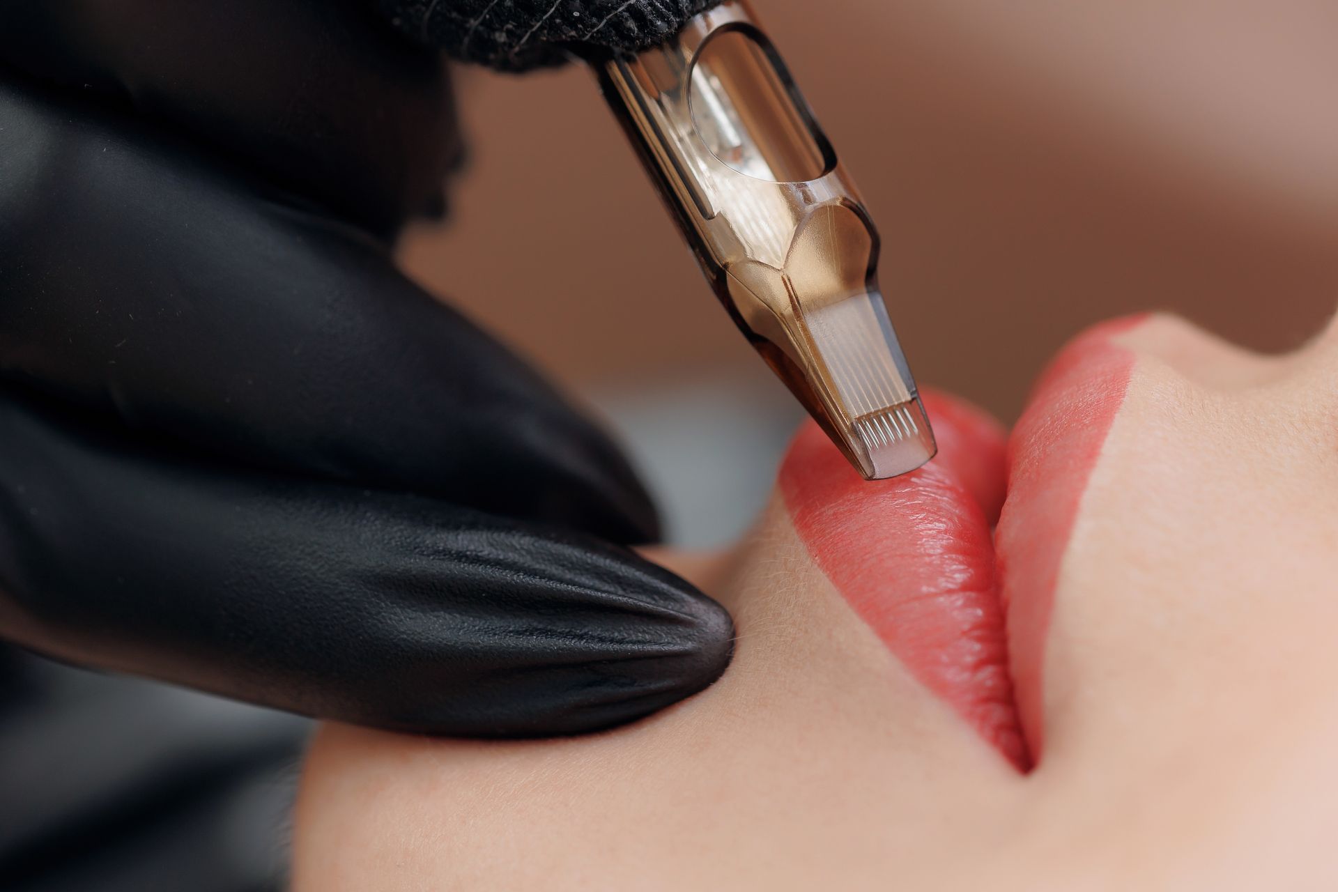 Lip blushing procedure: gloved hand using a tattoo pen on a person's lip, applying red pigment.