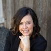 A woman in a black jacket is smiling with her hand on her chin.