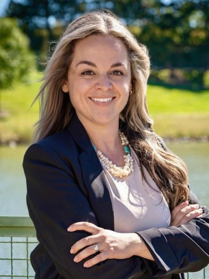 Woman in blazer smiles, arms crossed; background is greenery and water.