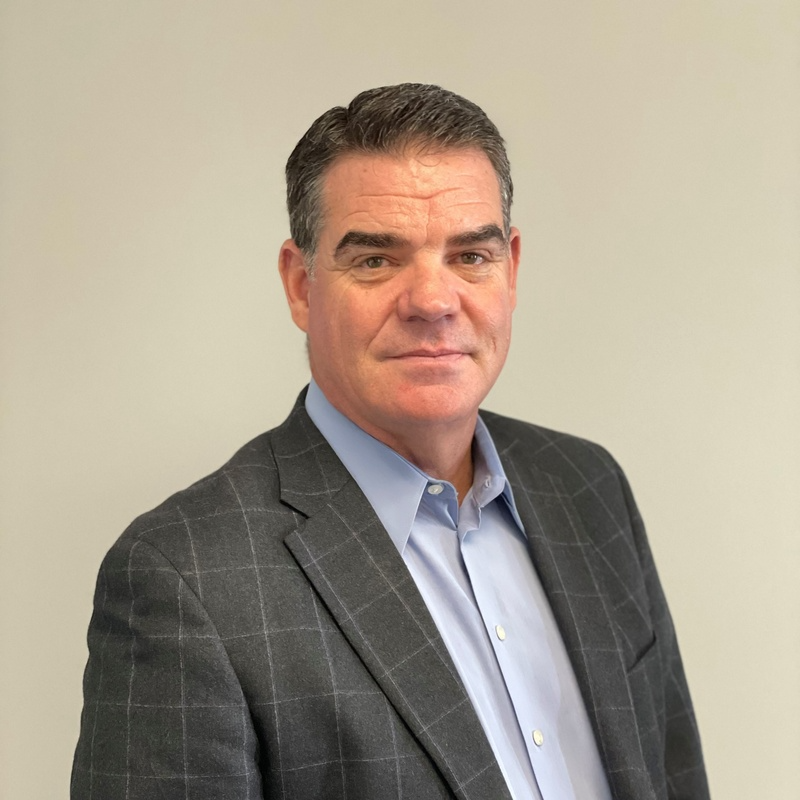 A man in a suit and blue shirt is standing in front of a white wall.