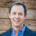 A man in a suit and blue shirt is smiling in front of a wooden wall.