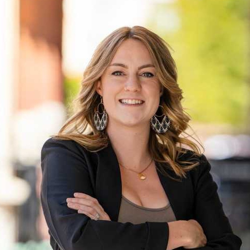 A woman in a black jacket and earrings is smiling with her arms crossed.