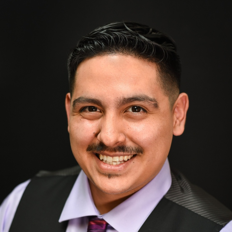 A man in a purple shirt and black vest is smiling for the camera.
