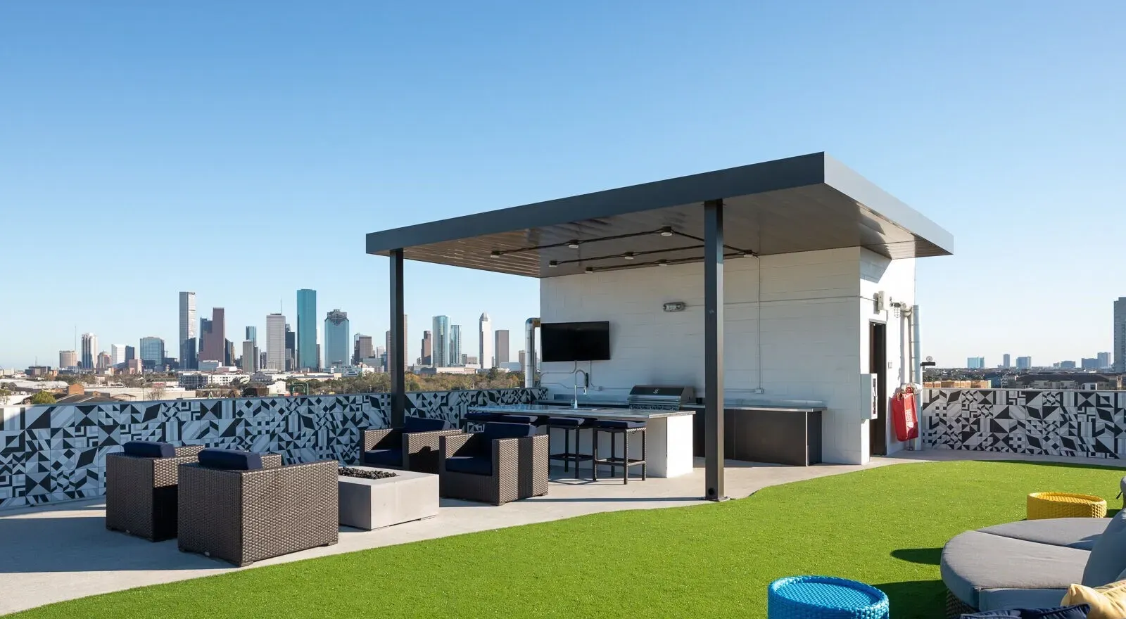 Rooftop lounge with modern seating and a small pavilion, overlooking a city skyline under a clear blue sky