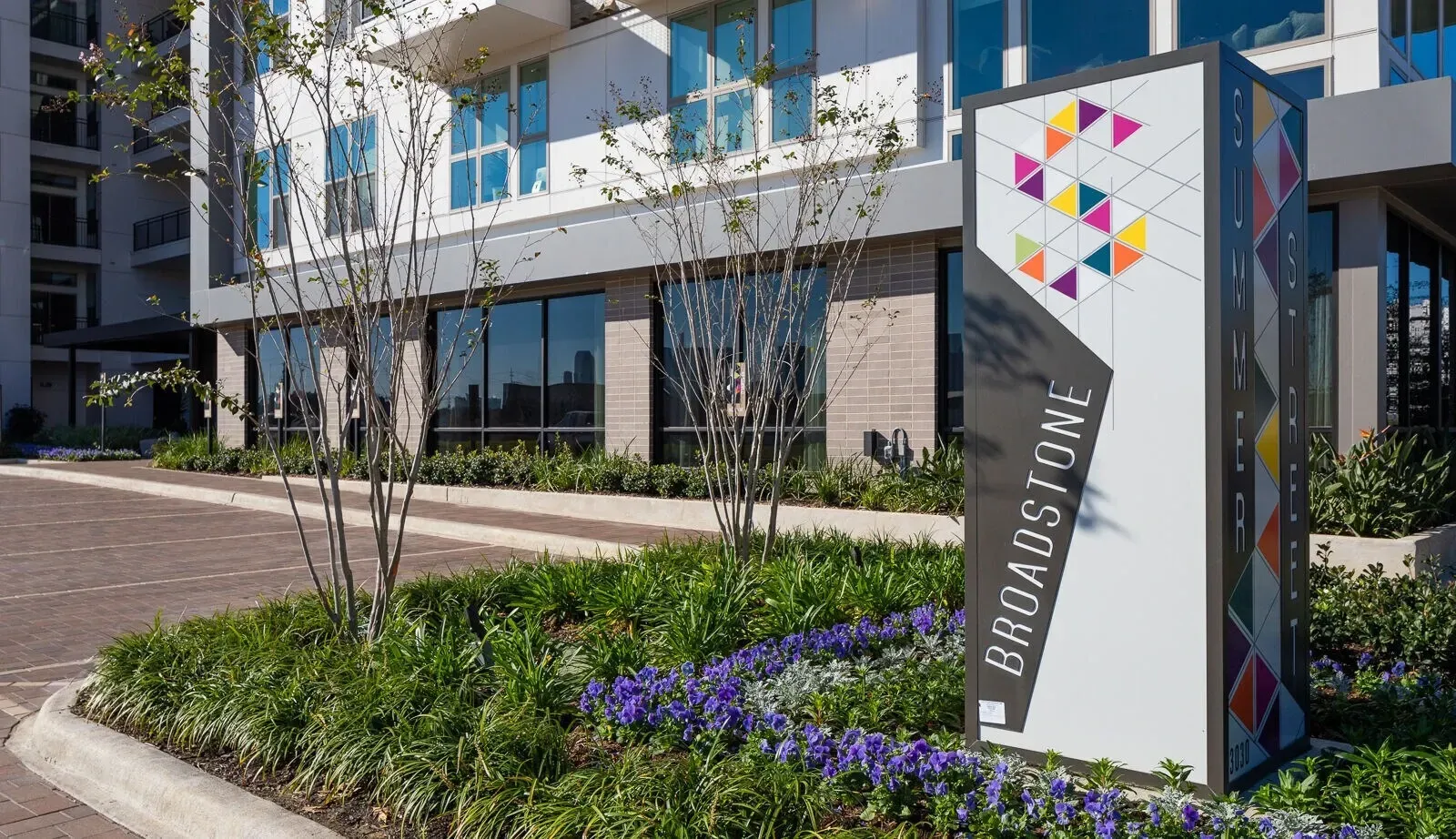 Modern apartment building entrance with a white sign, purple flowers, and landscaped walkway