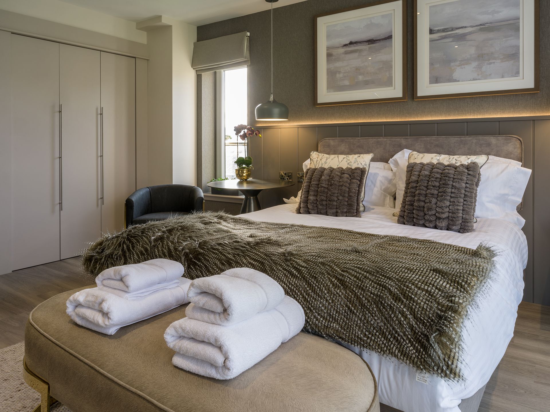 A bedroom with a bed , ottoman , chair and paintings on the wall.