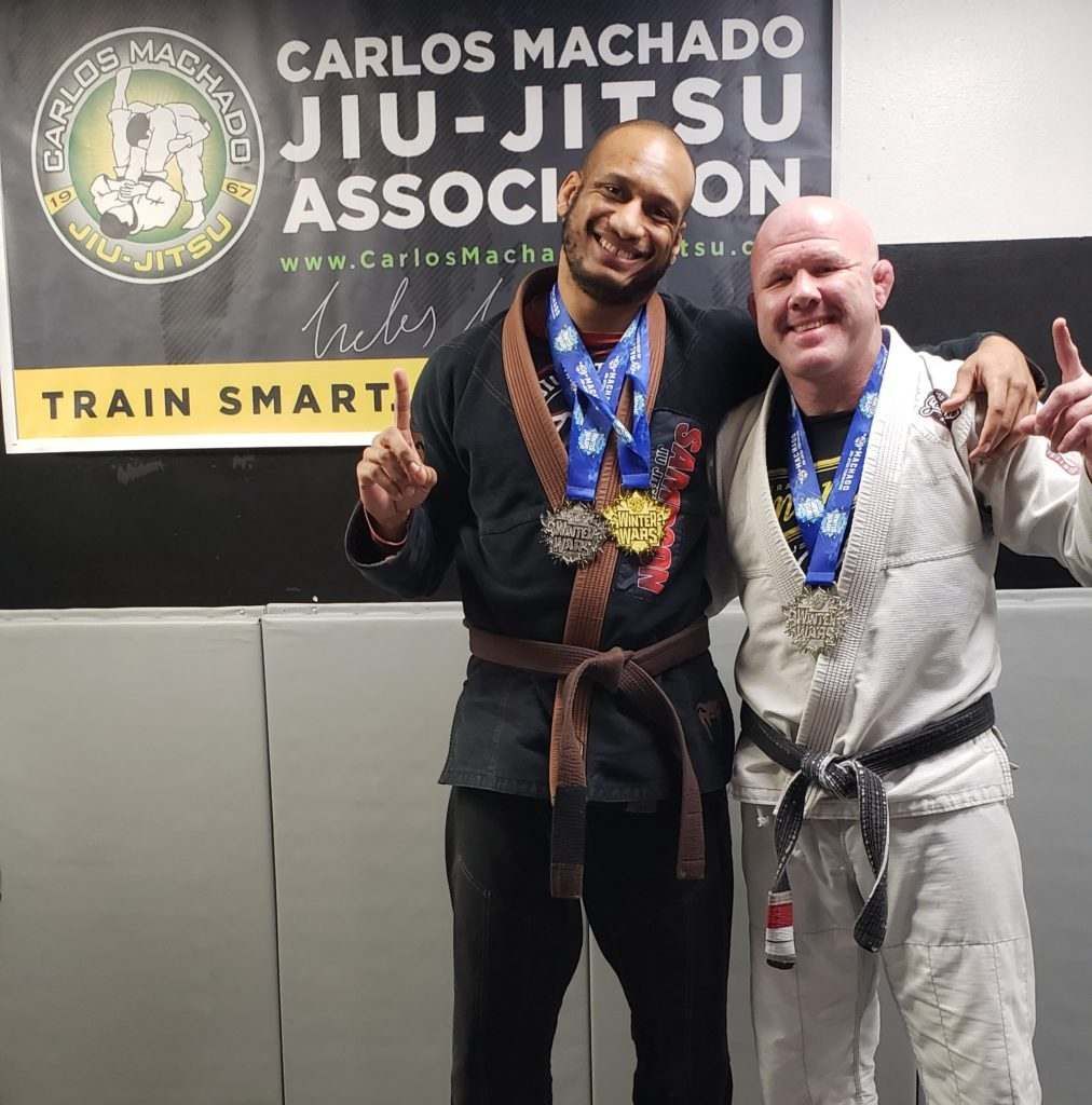 Two men are posing for a picture in front of a sign that says carlos machado jiu-jitsu association