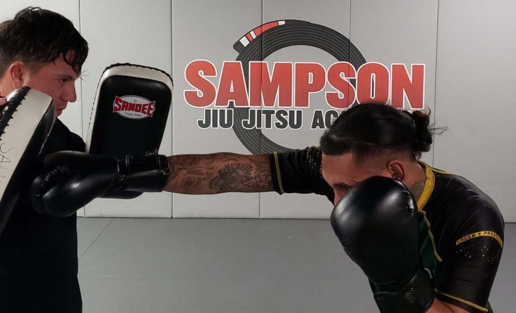Two men are boxing in a gym with sampson jiu jitsu academy in the background.