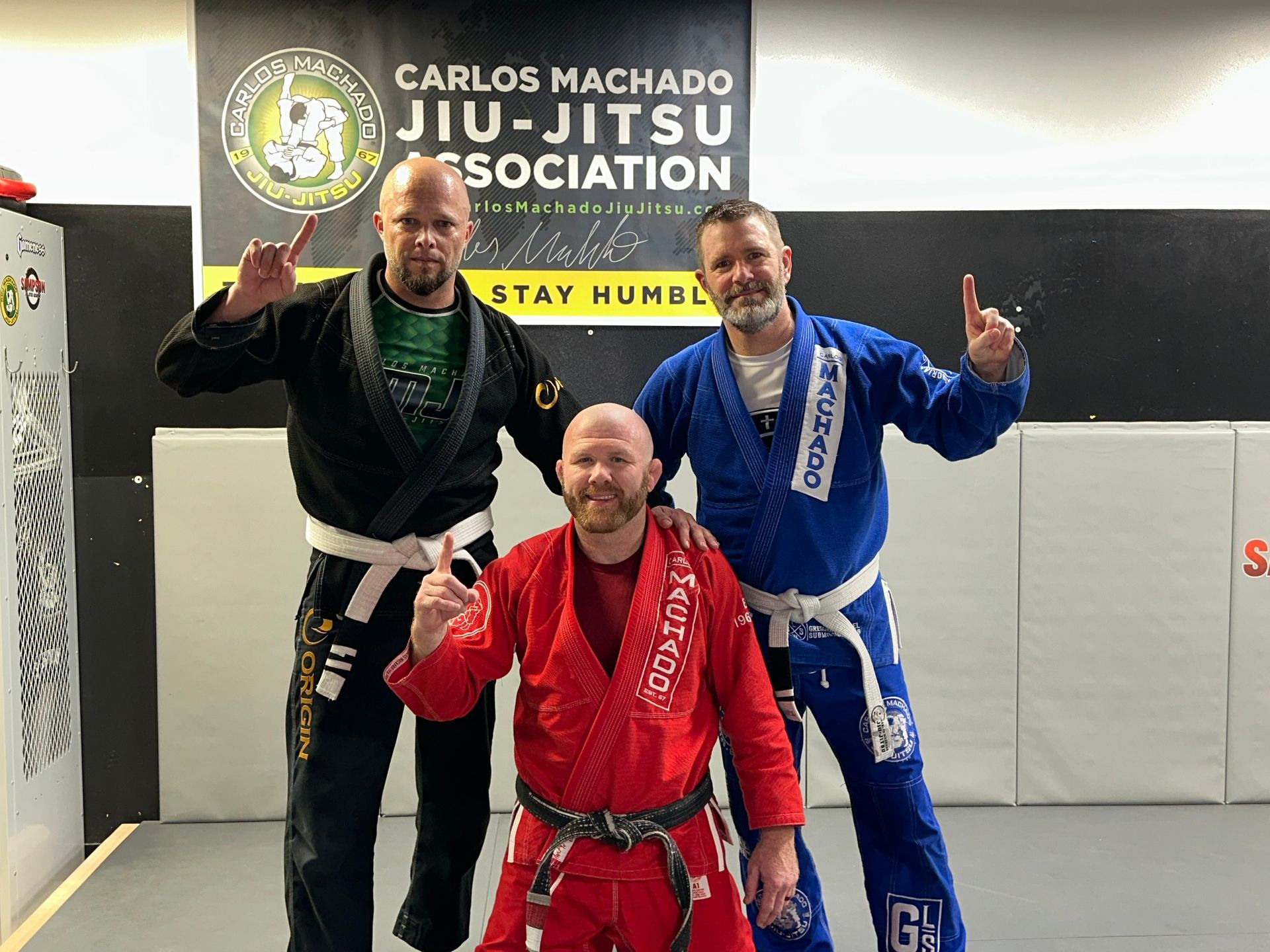 Three men are posing for a picture in front of a sign that says carlos machado jiu-jitsu association.