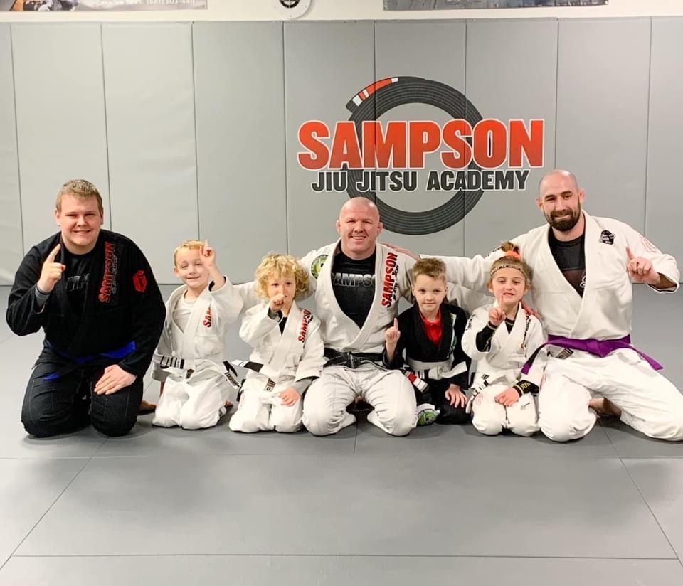 A group of men and children are posing for a picture at sampson jiu jitsu academy.