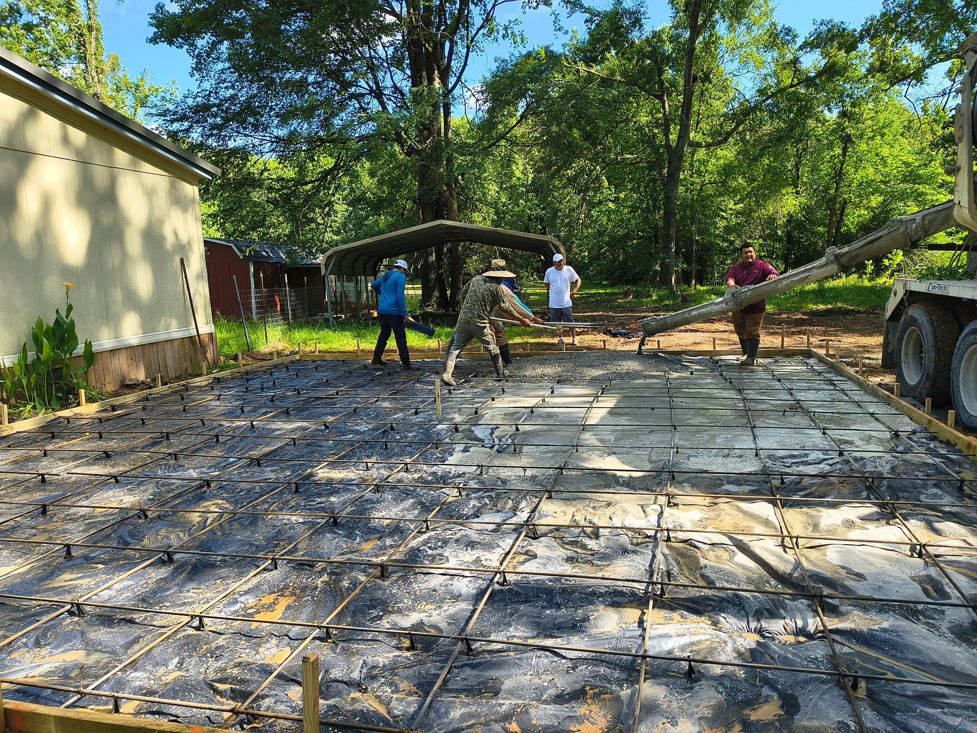 Workers pouring concrete for a patio. Rebar grid visible. Green trees in the background.